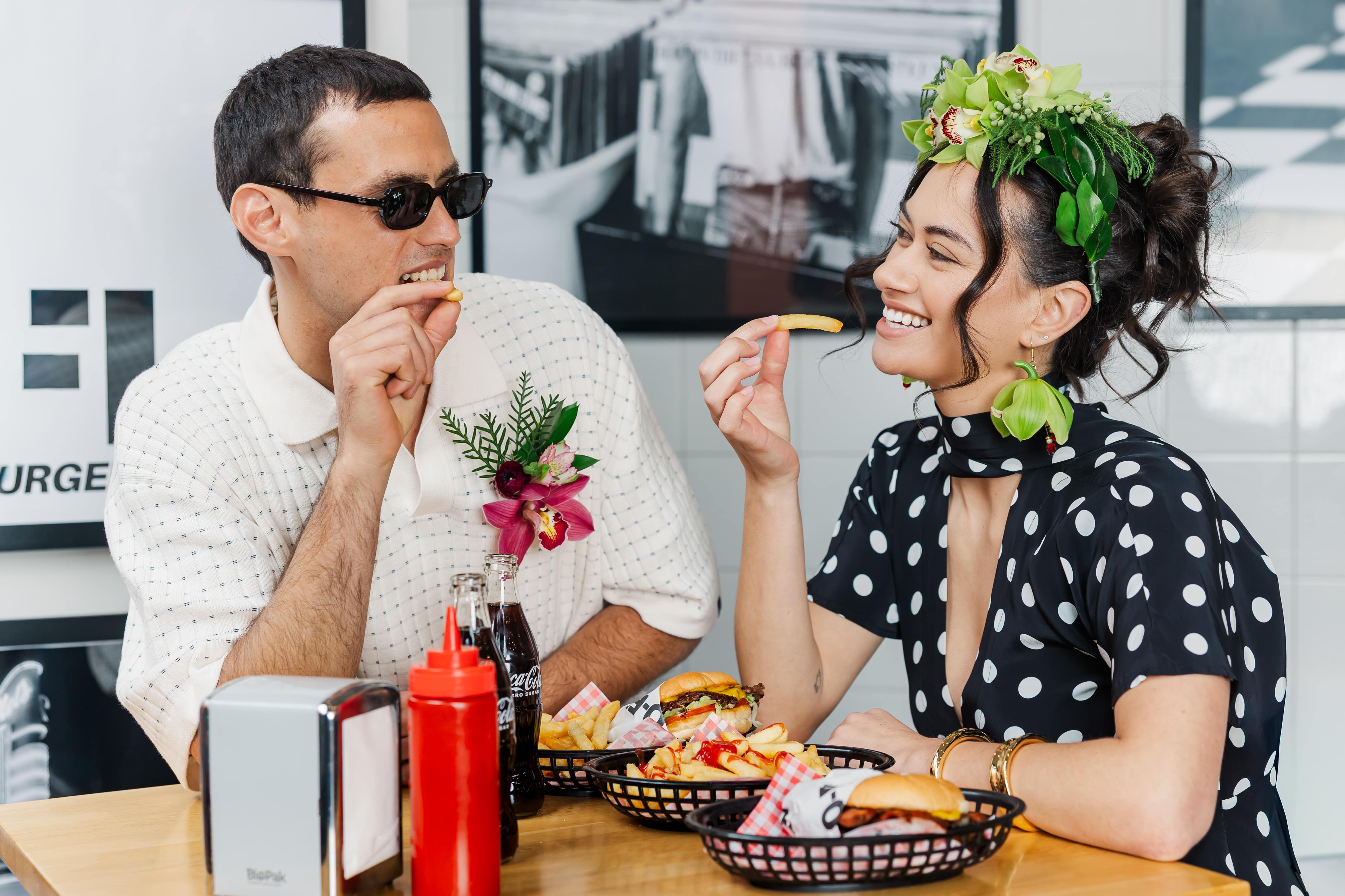 Amber enjoys a smash burger at Lo-Fi in The Crossing wearing a RUBY Gloria scarf dress, Andrea Biani Jules heels, Witchery cuff and bangle. Natural earrings and headdress by Thoisle & Co. Juan wears an I Love Ugly polo and Sunglasses Hut Ray Bans. Natural Boutonniere by Thoisle & Co.